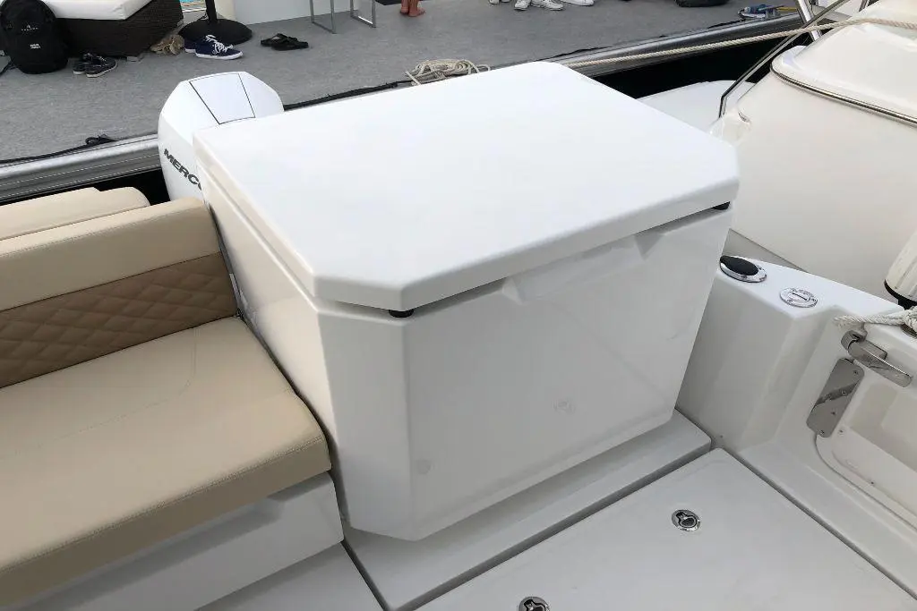 White cooler positioned on a boat beside a cream upholstered seat, with a grey deck and blurred figures in the background.